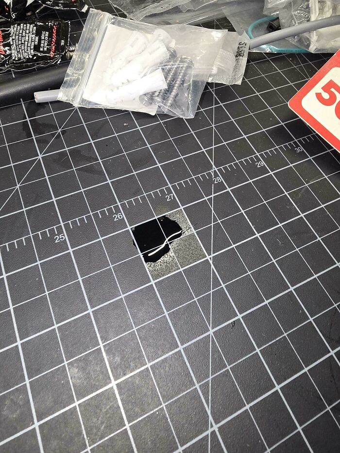 Black liquid spilled on a cutting mat with grid lines, next to plastic bags containing screws and wall anchors.