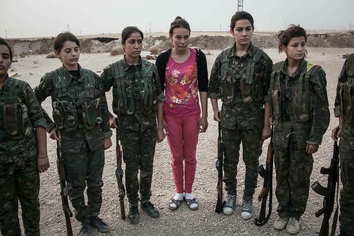 Group of young women wearing military uniforms standing next to one girl not in uniform in a desert setting.