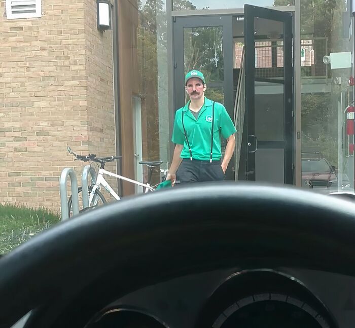 Man wearing a green uniform with suspenders and a cap stands outside near a bike rack and glass door, viewed from inside a car.