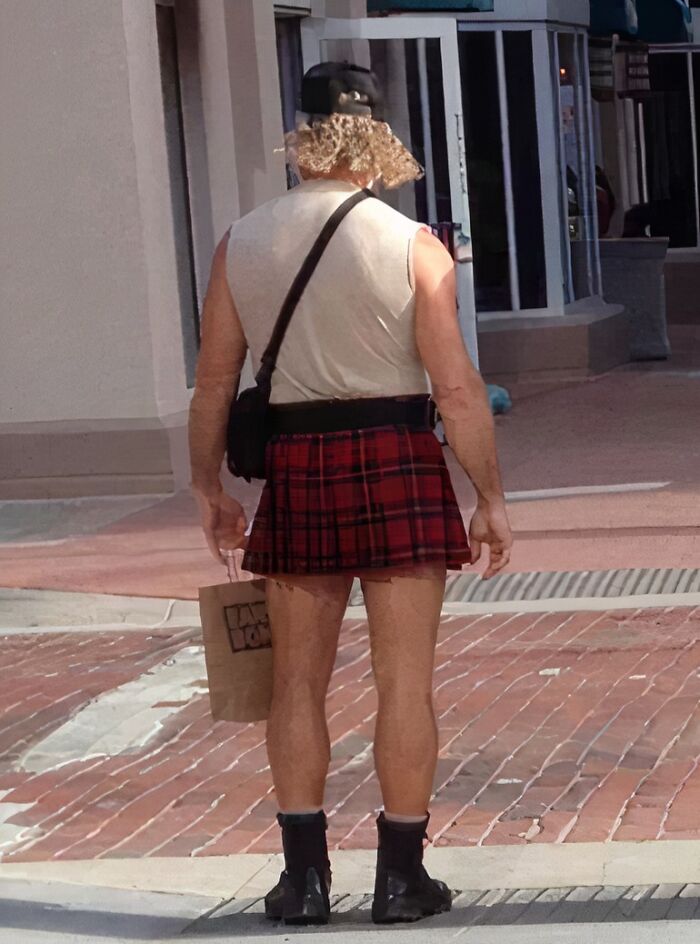 Man wearing an unconventional uniform with a red plaid kilt and a sleeveless shirt, standing on a sidewalk.