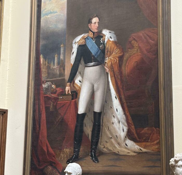 Portrait of a man wearing a royal uniform with a decorative cape, standing beside a throne in an ornate room.