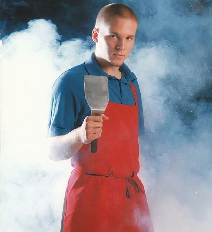 Young man wearing a funny uniform with a red apron and holding a spatula, surrounded by smoke or steam.