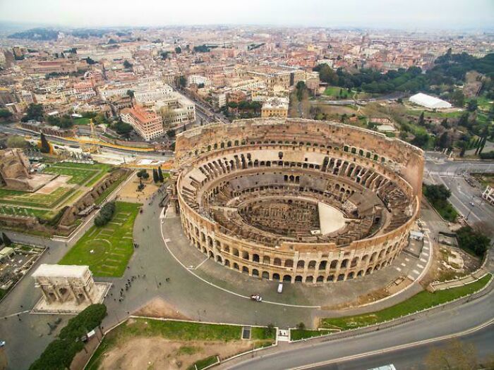 Aerial view of the Colosseum in Rome surrounded by urban buildings and roads in a dense city setting.