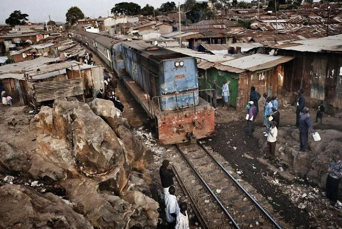 Rusty train passing through a crowded urban slum with makeshift homes and people standing near the tracks in an urban hell setting.