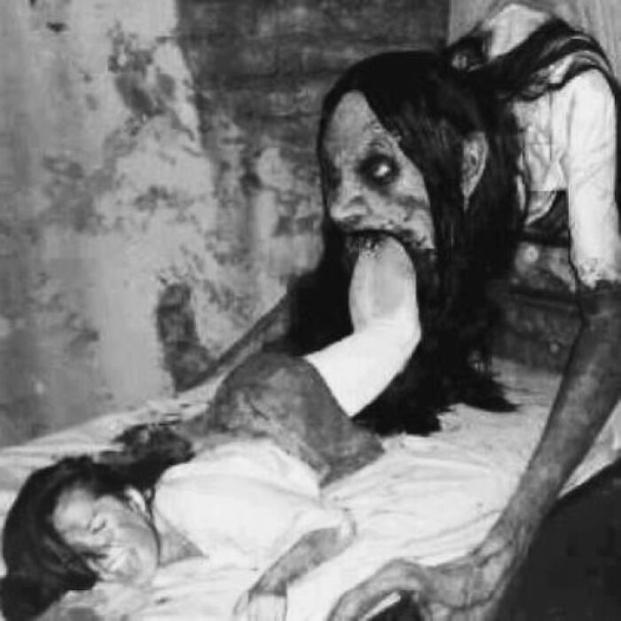 Black and white image of a terrifying figure with hollow eyes biting a person’s foot in a creepy setting.