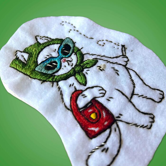 A charming embroidery creation of a cat with a green hat and blue sunglasses, carrying a red purse, inspiring embroidery hobbyists.