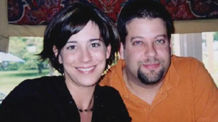 Smiling couple indoors, a woman with short dark hair and a man with a goatee in an orange shirt, captured in a terrifying photo.