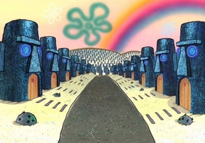 Rows of Easter Island-style stone houses with windows and doors along a sandy path under a colorful sky, urban hell echo chamber.