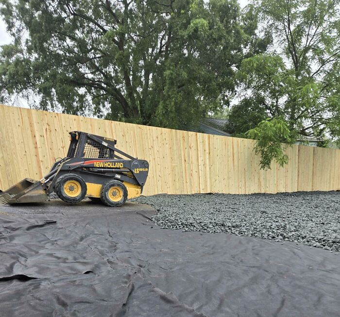 Small construction vehicle parked on black tarp and gravel near new wooden fence in a yard, showing landlord property changes.