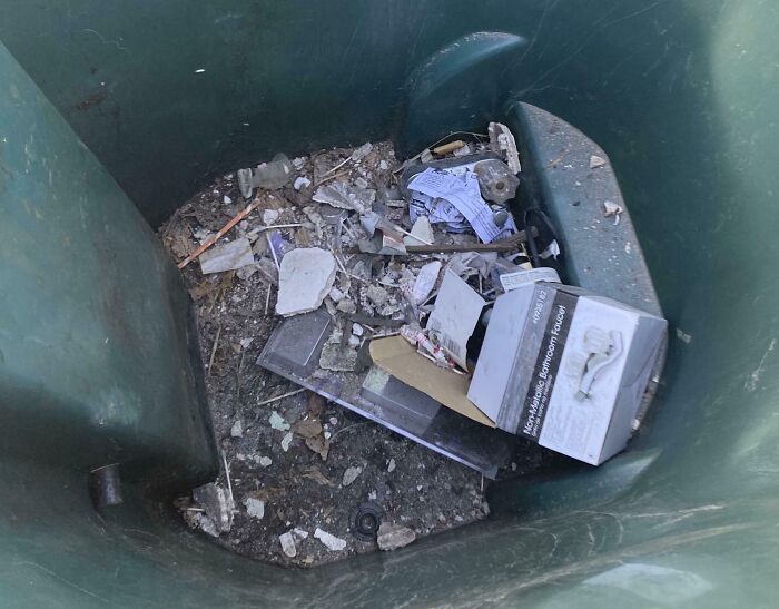 Trash and broken bathroom fixture parts dumped inside a large green container showing landlord neglect and poor rental conditions.