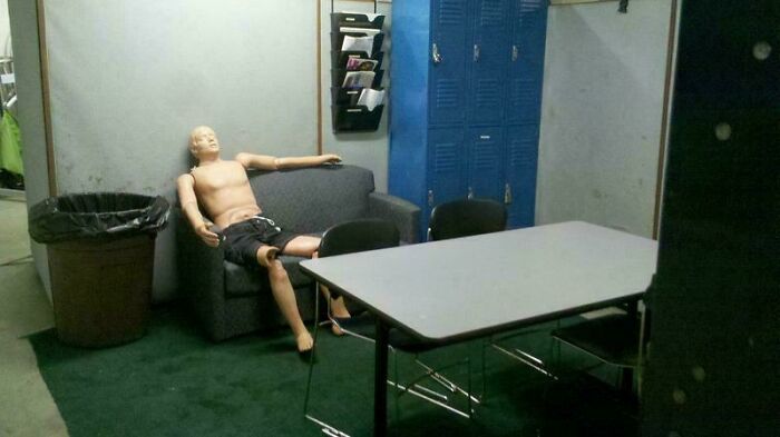 Break room resembling a prison with bare walls, metal lockers, a trash can, and a mannequin sitting on a worn couch.