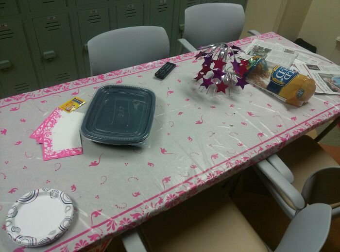 Break room with plastic-covered table, disposable plates, old chairs, and lockers in a prison-like environment.