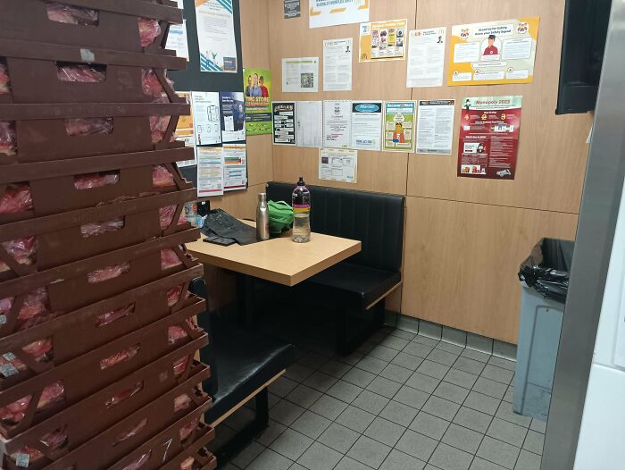 Cramped break room with worn black seating, wood-paneled walls covered in notices, and stacks of packaged goods on shelves.