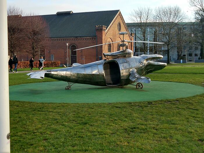 Metal dolphin-shaped sculpture resembling a Danish prison cell on green lawn near historic brick buildings in Denmark.