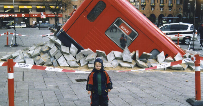 A Danish prison cell sculpture tilted in a public square surrounded by concrete blocks and a child standing nearby.