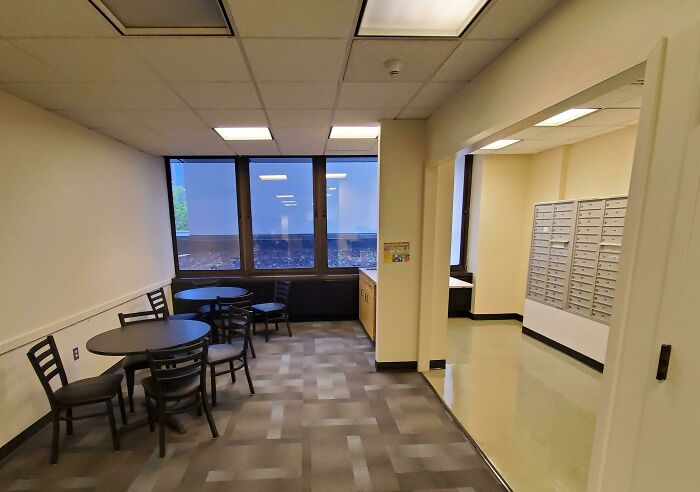 Sparse break room with basic tables and chairs, dim lighting, and a dull atmosphere resembling a prison setting.