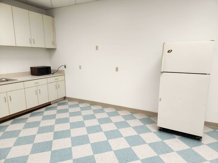 Sparse break room with white cabinets, old refrigerator, microwave, and checkered blue and white floor tiles.