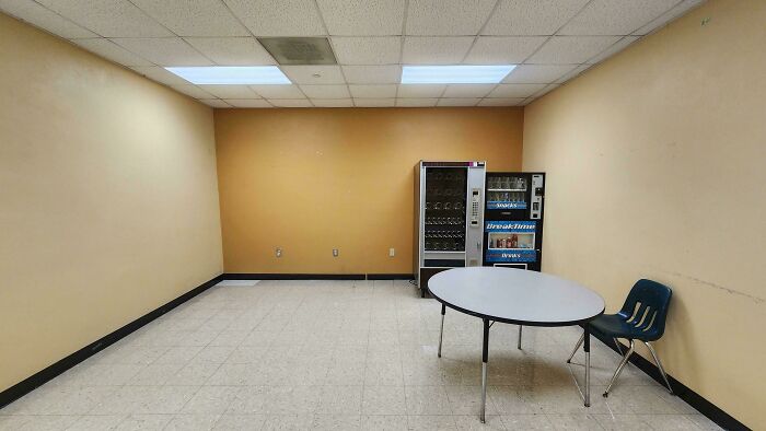 Sparse break room with old vending machines, a round table, and a single chair, resembling a prison environment.