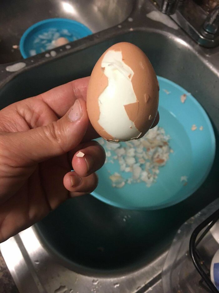 Hand holding a partially peeled boiled egg over a kitchen sink with a bowl of eggshells, everyday things humbling adults.