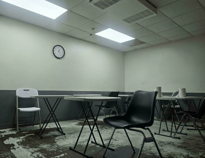 Bleak break room with c*****d floors, dingy walls, sparse furniture, and harsh fluorescent lighting resembling a prison setting.