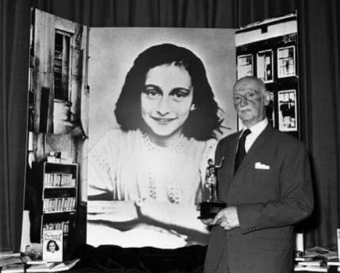 Black and white photo of an older man holding a trophy standing beside a large portrait in a perception of time comparison display.