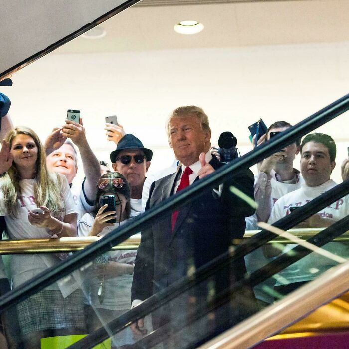 Donald Trump giving thumbs up on an escalator surrounded by a crowd capturing the moment with phones, highlighting time perception.