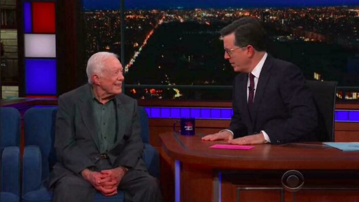 Two men having a conversation on a late-night talk show set with a cityscape visible through the window behind them.