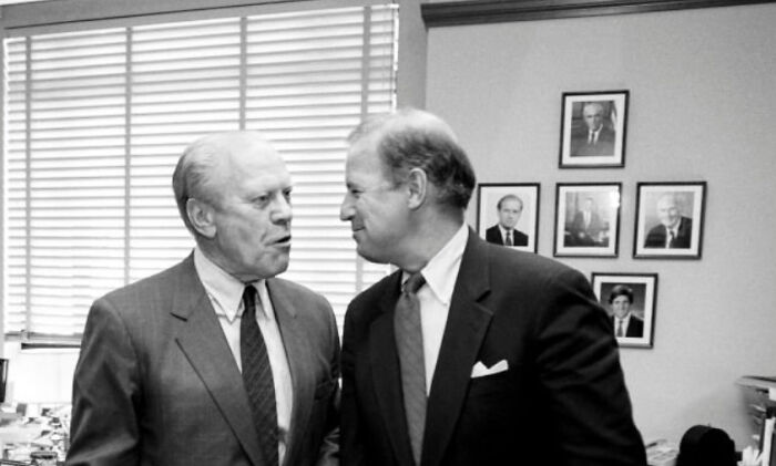 Two men in suits conversing in an office with framed portraits on the wall, illustrating time perception comparisons.