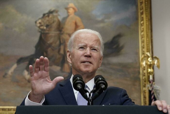 A man in a suit speaking at a podium with microphones in front of a historical painting background.