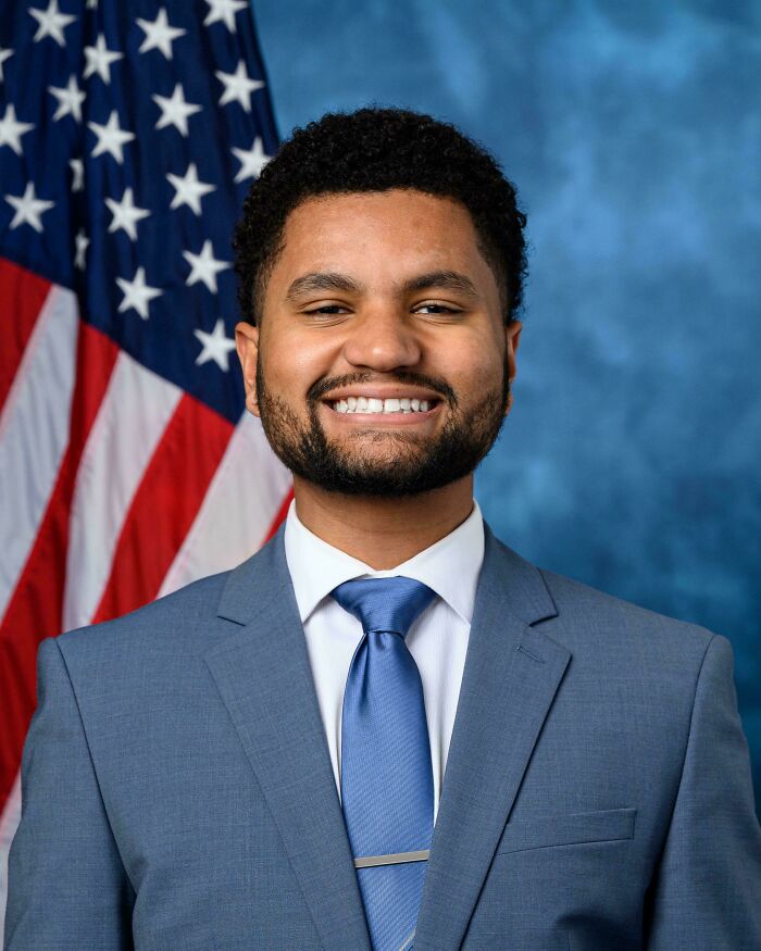 Smiling man in blue suit and tie standing in front of American flag illustrating perception of time concept.