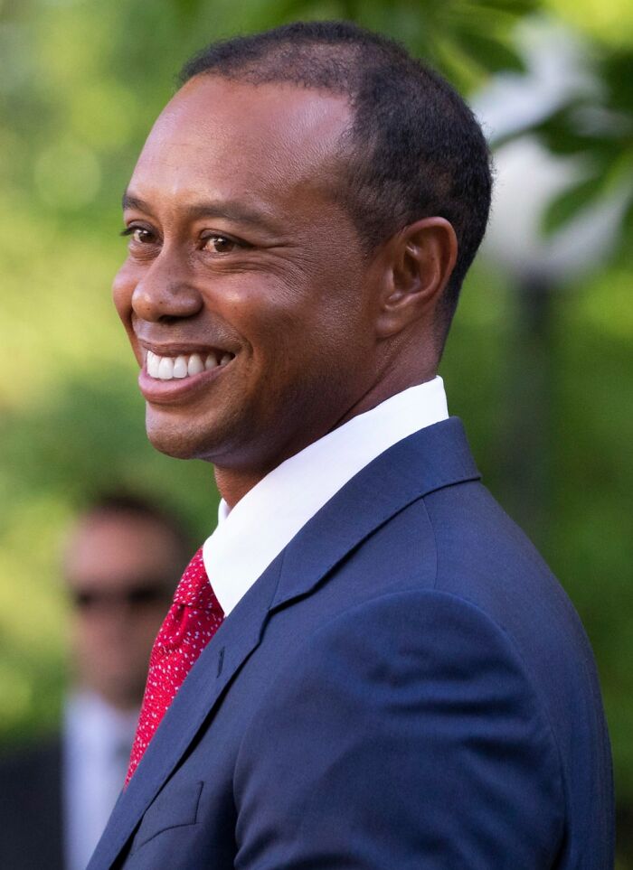Smiling highest paid athlete in 2025 wearing a navy suit and red tie outdoors with a blurred green background.