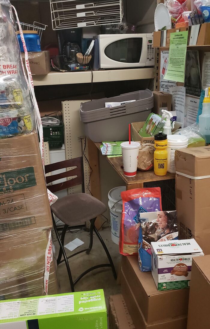 Cluttered and cramped break room with boxes, a small chair, microwave, coffee maker, and various supplies creating a prison-like feel.