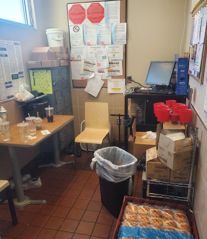 Cramped break room resembling a prison, cluttered with boxes, lockers, a single chair, and a small table with empty cups.