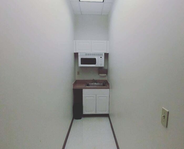 Narrow break room with minimal kitchen amenities, resembling a prison cell with plain walls and a basic sink area.