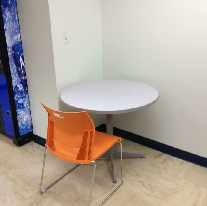 A small, stark break room with a single orange chair and round table beside a vending machine, resembling a prison break room.