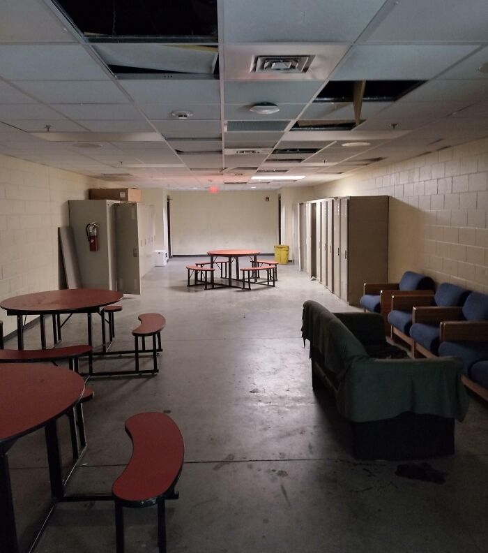 Dull and worn break room with damaged ceiling, basic benches, lockers, and sparse seating resembling a prison environment.