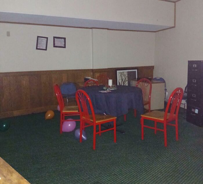 Dimly lit break room with red chairs around a table, scattered balloons, and a dull prison-like atmosphere.