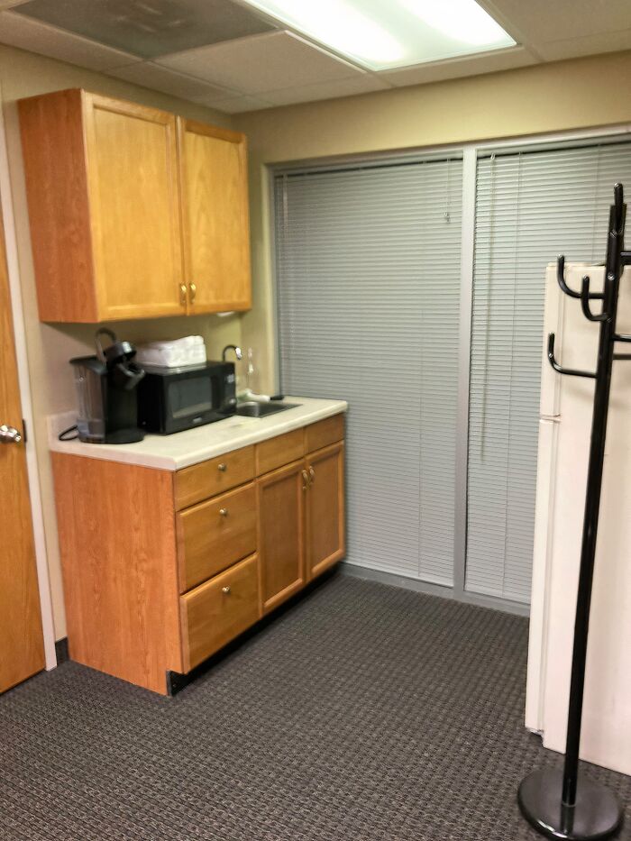 Small office break room with wooden cabinets, microwave, coffee maker, blinds, and carpet, showing worst break rooms ever design.