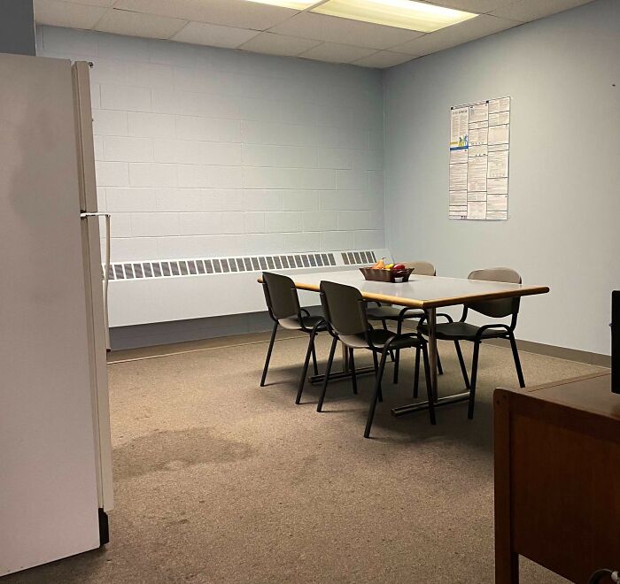 A bleak, prison-like break room with a simple table, four chairs, a bench heater, and a plain wall with posted papers.