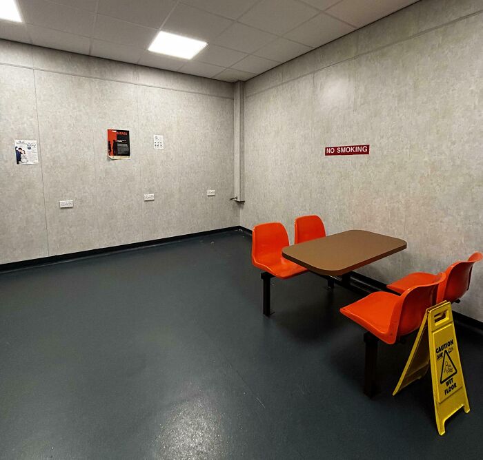 Sparse break room with prison-like gray walls, orange attached chairs, a table, and a caution wet floor sign.