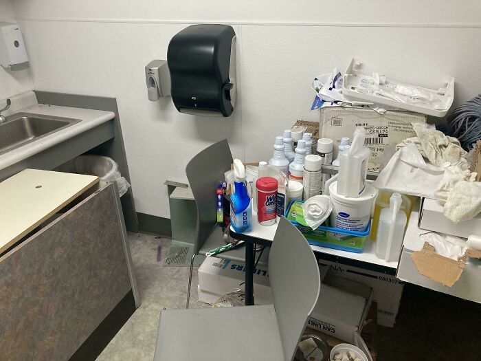 Cluttered and unkempt break room with cleaning supplies and boxes, resembling a prison-like environment.
