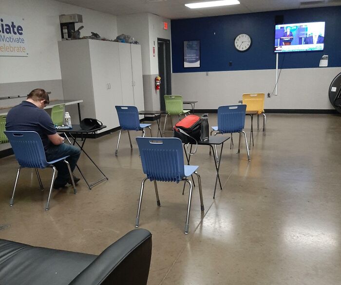 Sparse break room with mismatched chairs and desks, a TV mounted on the wall, and a person sitting alone.