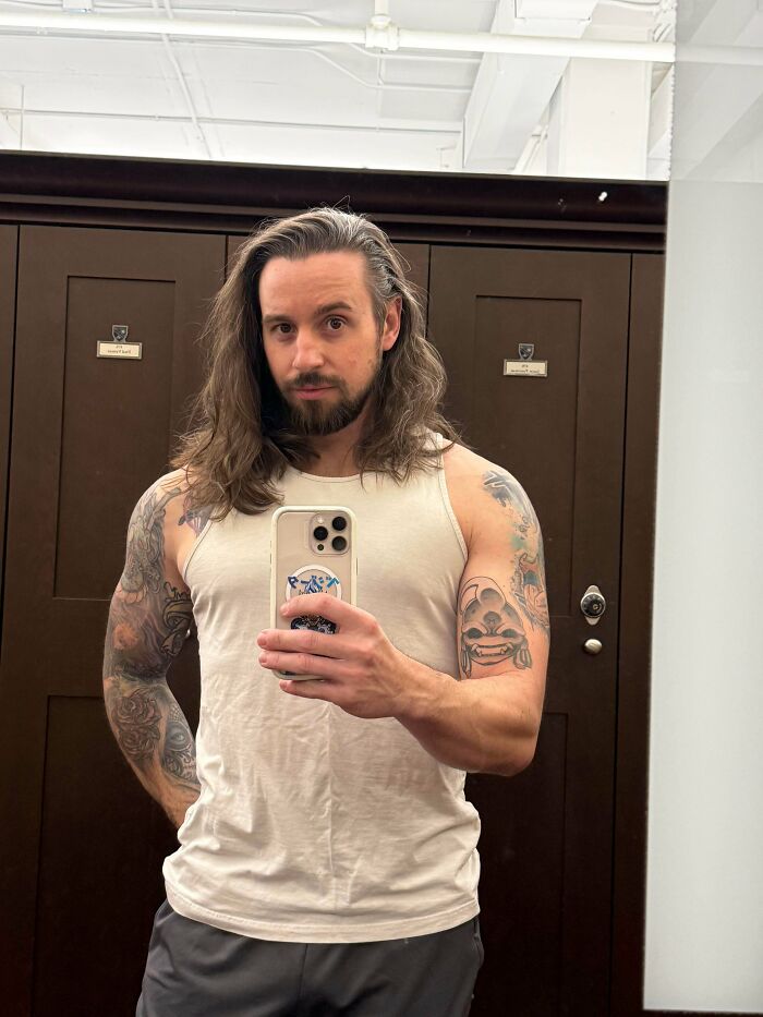 Man with fierceflow long hair and tattoos wearing a sleeveless shirt taking a mirror selfie in a locker room.