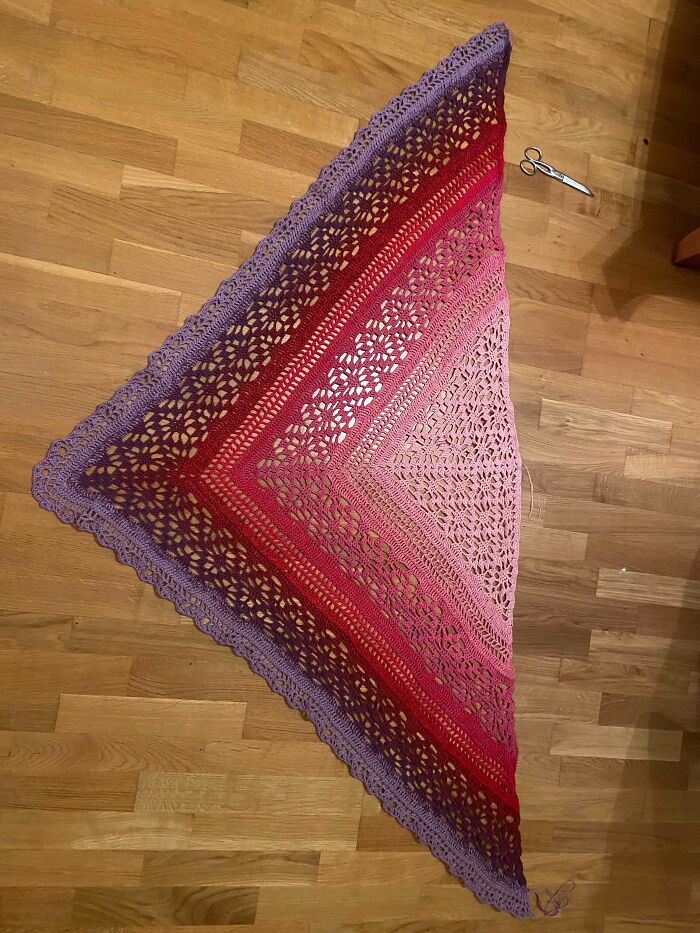 Crocheted triangular shawl with gradient colors from pink to purple laid on wooden floor next to small scissors.