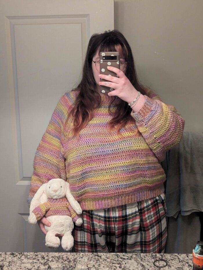 Person wearing a colorful crocheted sweater holding a matching crocheted sweater on a stuffed rabbit in a bathroom.