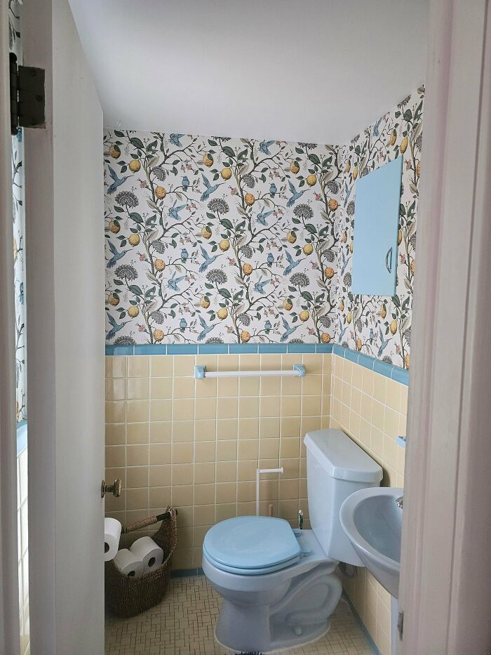 Bathroom home decoration with floral wallpaper, light blue and yellow tiles, and a wicker basket with toilet paper rolls.