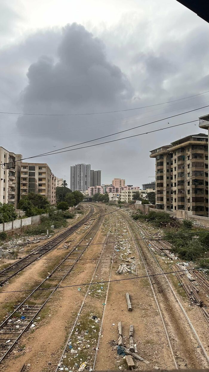 Urban hell scene showing abandoned train tracks covered in trash, surrounded by neglected buildings under a cloudy sky.