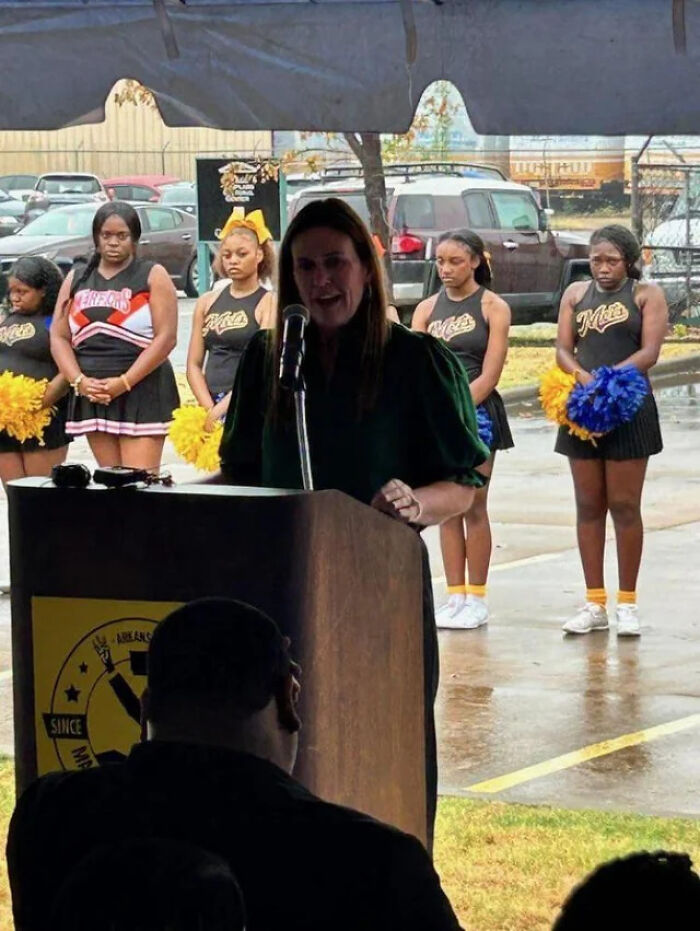 Woman speaking at podium during outdoor event with cheerleaders standing behind in rain, illustrating there was an attempt.