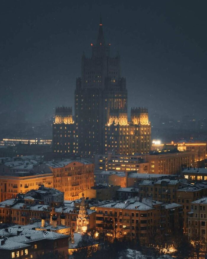 A towering evil building illuminated at night, surrounded by smaller city buildings under a dark, snowy sky.