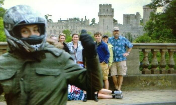 Person wearing a helmet and jacket giving a thumbs up with a group of people and a castle in the background, perfect timing photo.